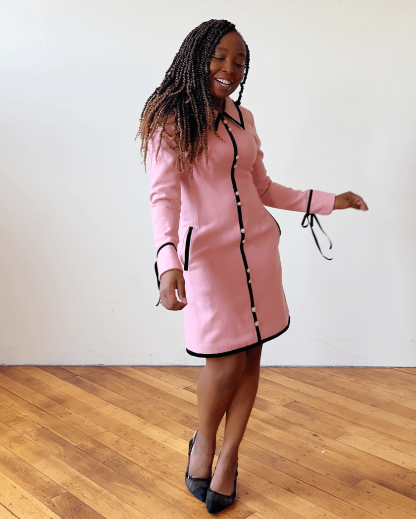 Woman wearing a pink wool dress with black trim, standing on a wooden floor.